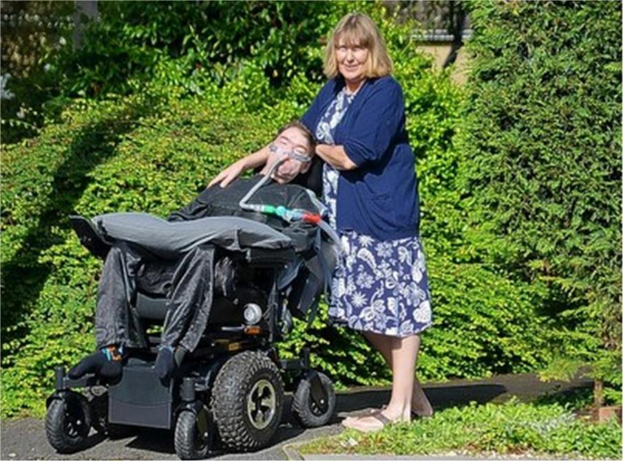 Daniel Baker in a motorised wheelchair with medical equipment, accompanied by a standing companion (his Mother) in an outdoor setting with greenery and a paved path; the scene conveys care, support, and companionship in a peaceful natural environment.