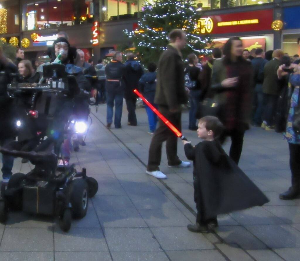 Me in my wheelchair dressed as Darth Vader with a two year old boy also dressed as Darth Vader attacking with his red light Saber