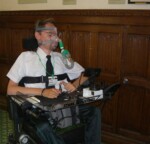 Mr Baker in his wheelchair sitting in front of a wood panelled wall inside parliament