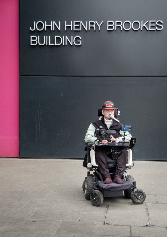 Daniel Baker in his wheelchair sitting in front of the John Henry Brookes building 