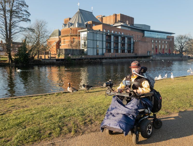 Daniel Baker in his wheelchair next to the River Avon, with the Shakespeare Royal Theatre across the river behind him