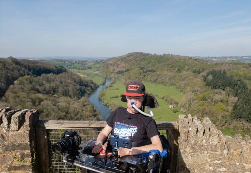 Easter Weekend - Daniel Baker in his wheelchair wearing a Guardians of the Galaxy T-Shirt at the top of Symonds Yat, with a view of hills and a winding river behind him