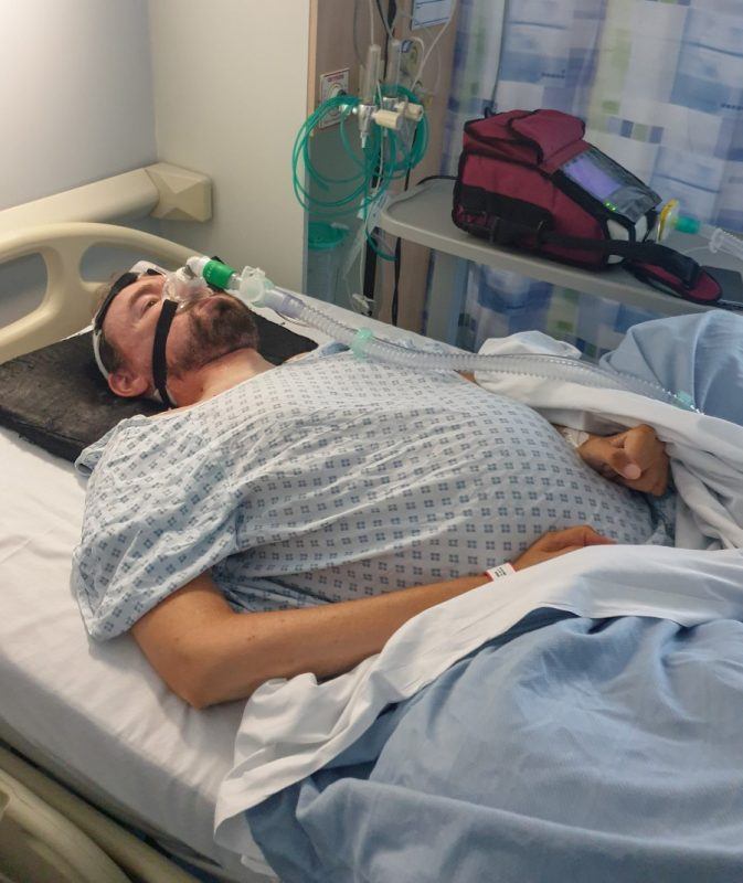 Daniel in a hospital gown laying in a hospital bed with his distended abdomen showing under his gown