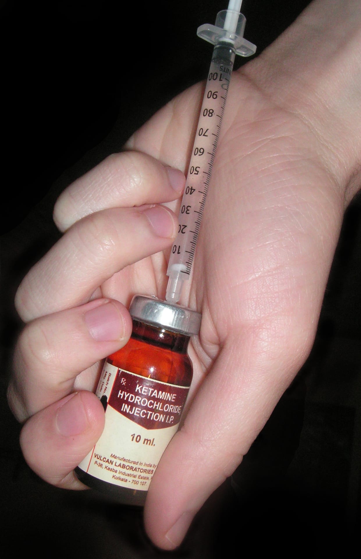 The image shows a close-up of a hand holding a small glass vial labeled Ketamine Hydrochloride Injection. This setup suggests a clinical or procedural context—perhaps medical preparation for anesthesia or pain relief, as ketamine is commonly used for those purposes.