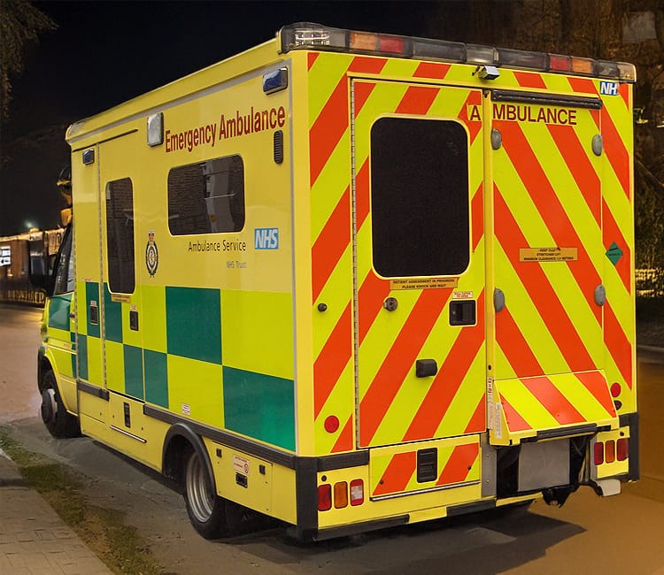 The image shows the rear view of a yellow emergency ambulance parked on a street in the UK at night. It's clearly part of the NHS fleet—green and yellow checkered markings run along the sides, while high-visibility red and yellow diagonal stripes cover the back doors. Prominent text reads “Emergency Ambulance,” and the NHS branding includes “Ambulance Service NHS Trust.”