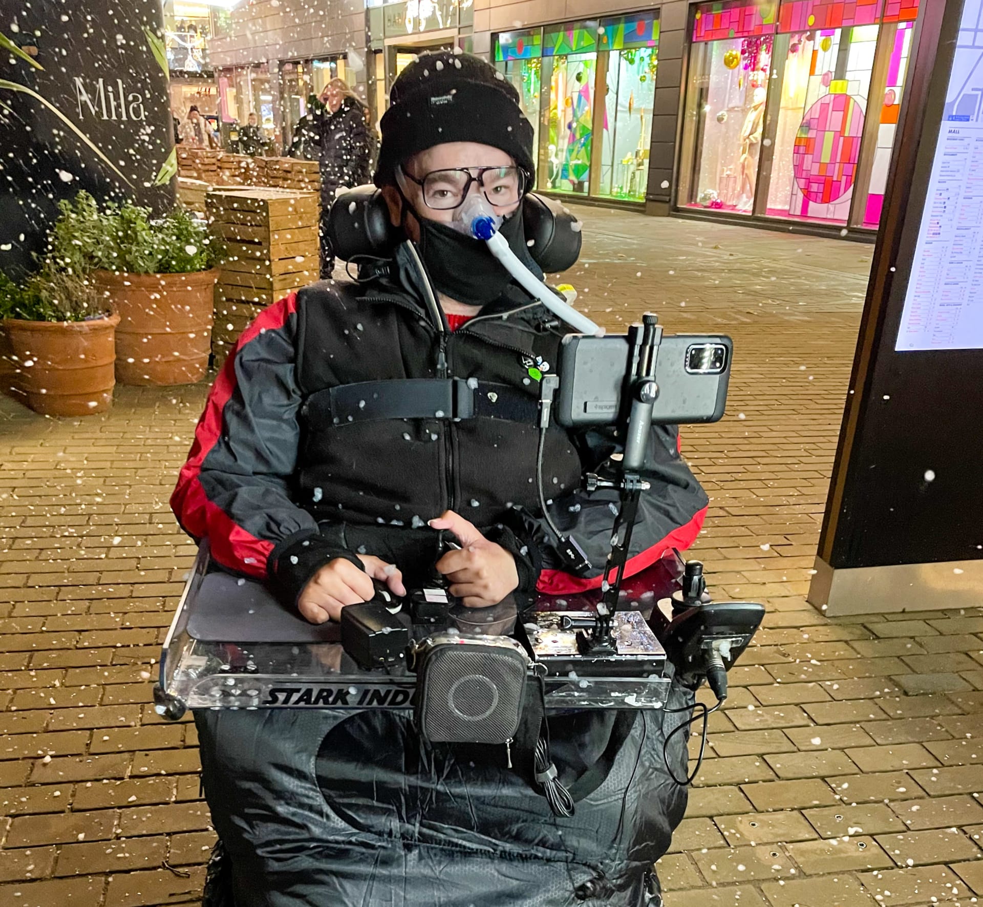 The image features a person in a motorized wheelchair that’s been outfitted with a full tech setup, there’s a front tray holding a mounted smartphone, speaker, and other electronic gear, suggesting a custom-built accessibility rig. They’re wearing a black and red jacket, and the setting is outdoors in a lively, retail area, with bright storefronts and signage adding plenty of visual interest. There’s also a festive vibe, artificial snow drifts through the air, and decorative lights twinkle in the background. What really stands out is the bold "STARK INDUSTRIES" label on the front tray, an unmistakable nod to the Marvel universe, as if Tony Stark himself might've engineered this mobility suite.