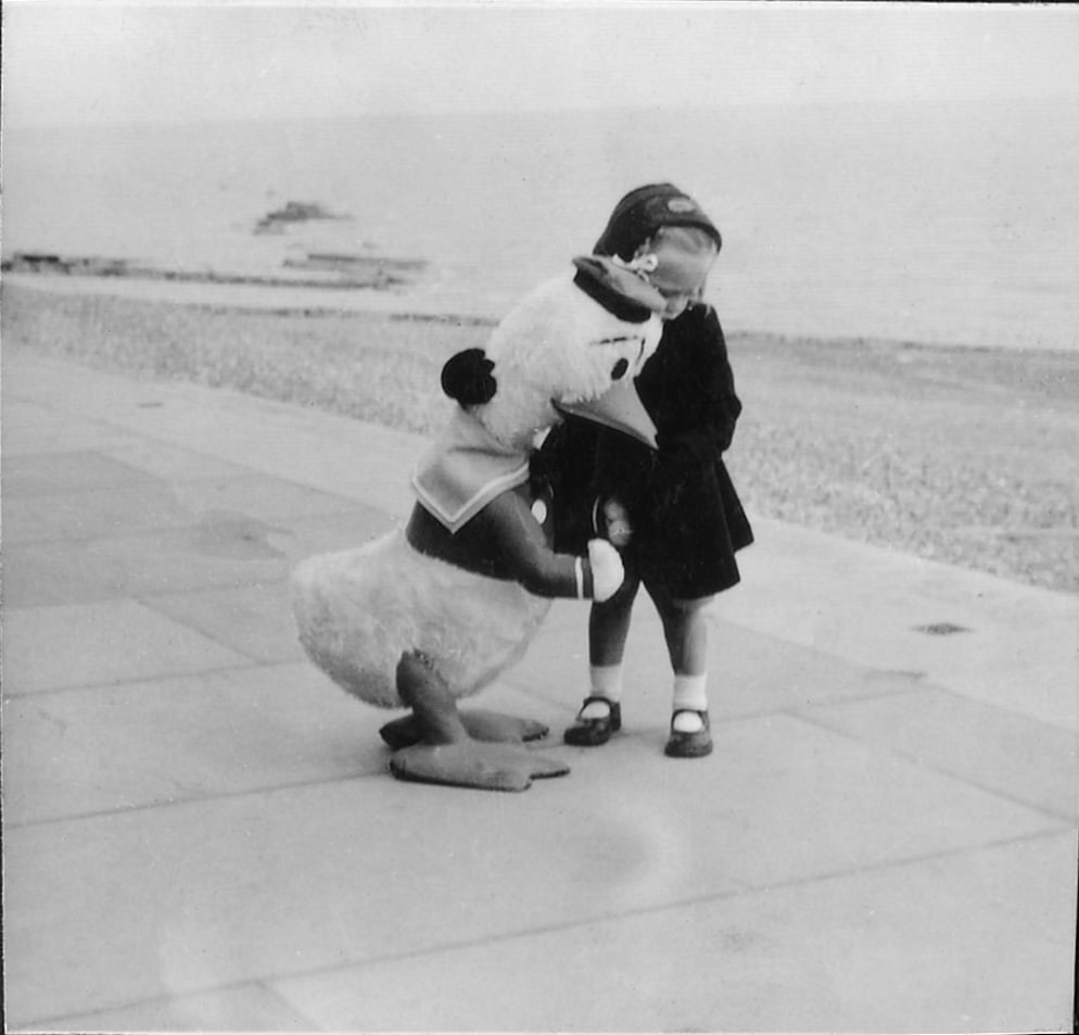 Black and white photo of a young girl walking down a seafront holding a Donald Duck stuffed toy