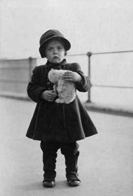 Black and White photo of a young girl with a hat standing on a seafront holding a toy