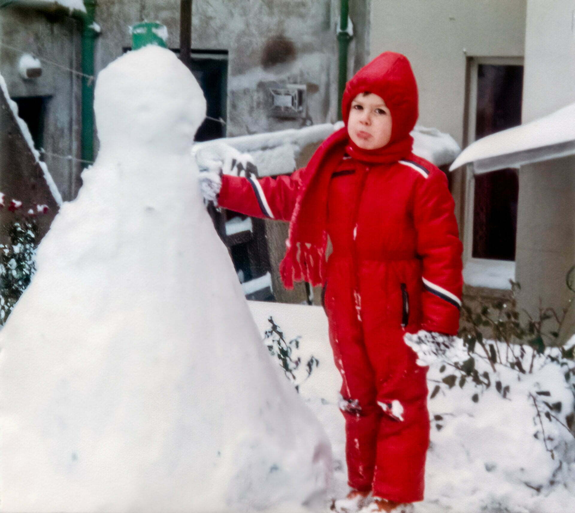 A young Daniel Baker in a red winter suit standing next to a snowman