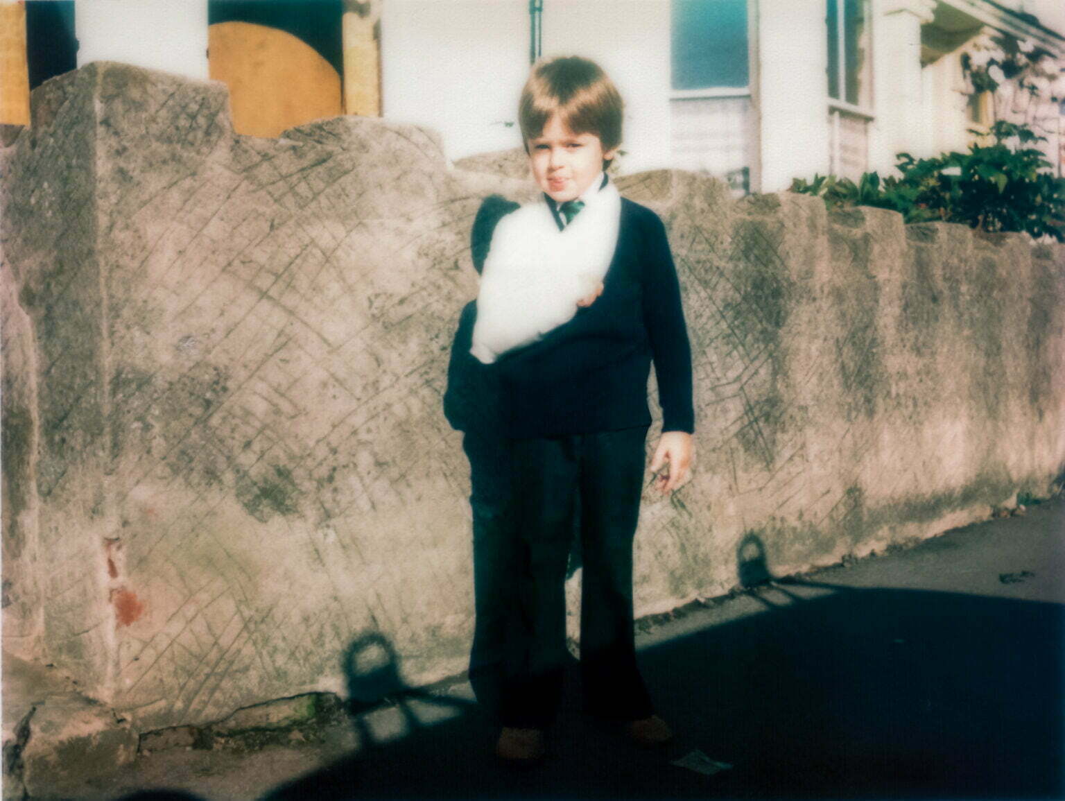 A young Daniel Baker standing next to a wall, wearing a school uniform with his arm in a sling