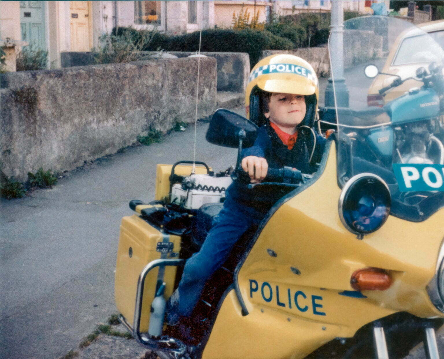 a young boy sitting on a yellow police motorcycle wearing a police helmet from the 1970's.