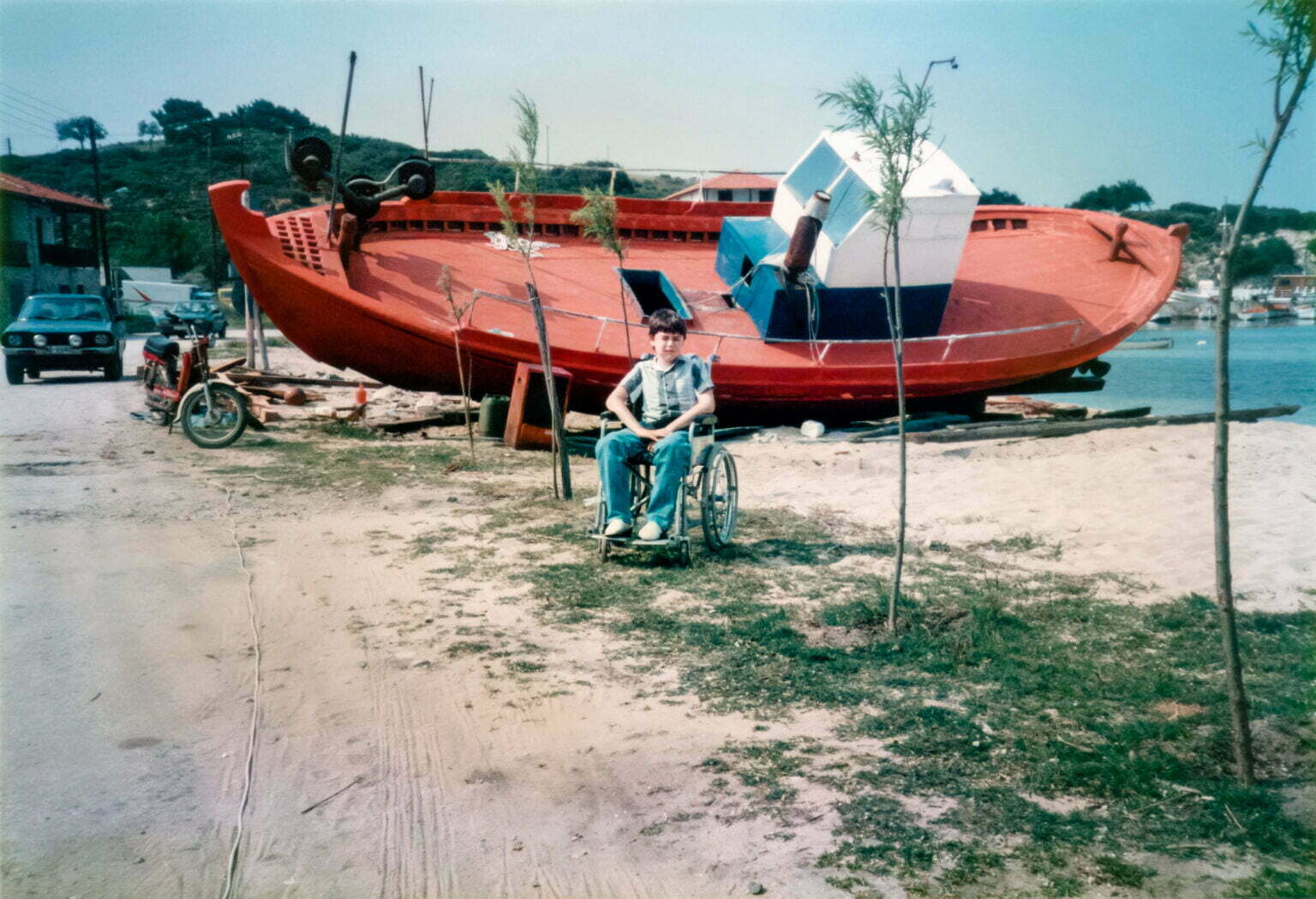 A young Daniel Baker in his wheelchair sitting in front of a red boat on a beach in Greece