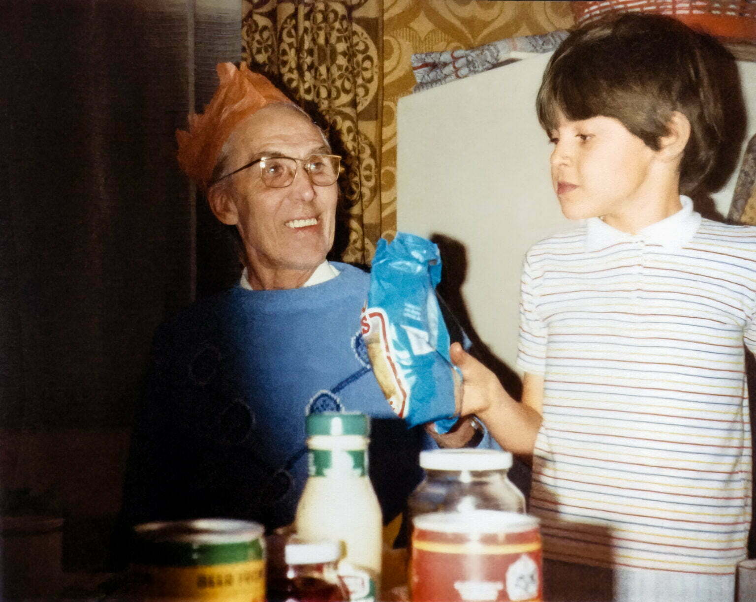 An older man wearing a paper Christmas hat smiling at a young boy (Daniel and his Granddad)