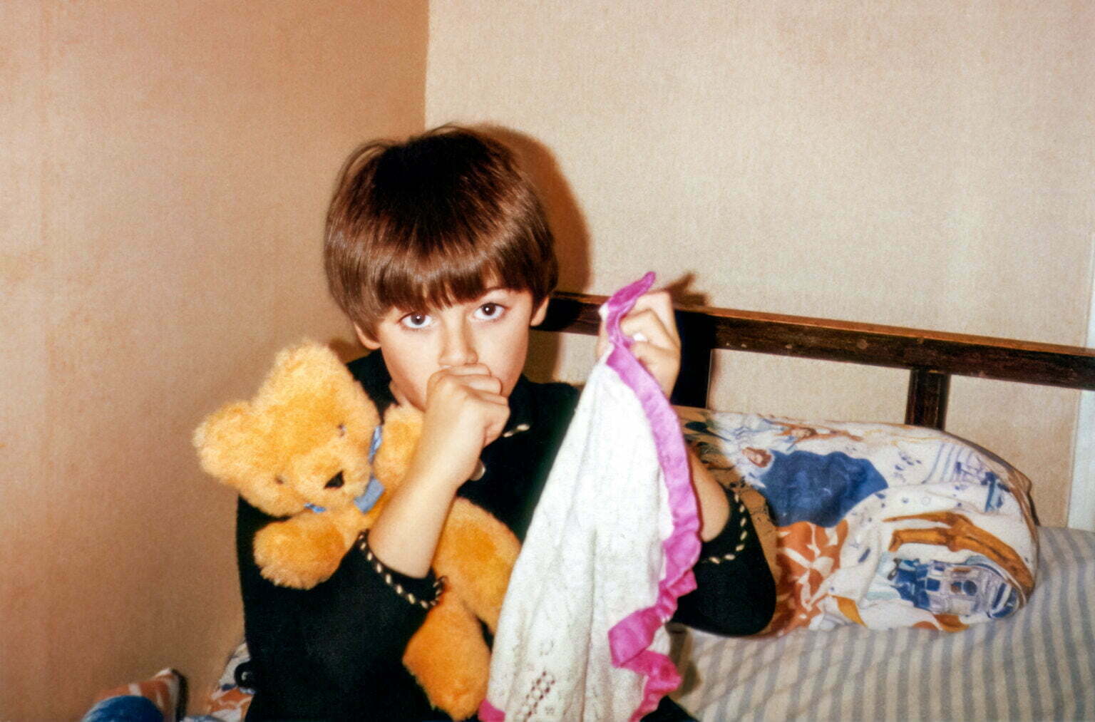 a young boy holding a teddy bear in his hand sitting on a bed with a Star Wars pillow.