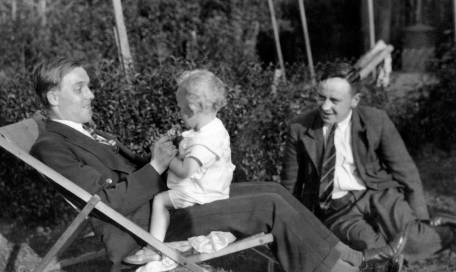 Black and white photo of a two men sitting in a garden, one is holding a young child