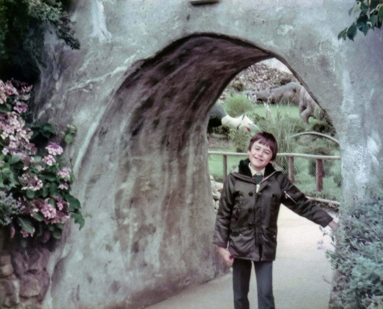 A young Daniel Baker standing in a stone tunnel with a dinosaur model behind him.