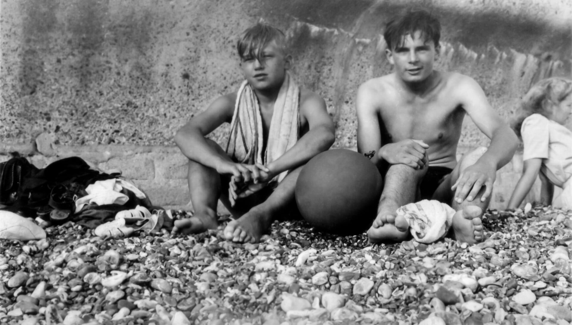 Black and white photo of two young men sitting on a pebbled beach with a ball