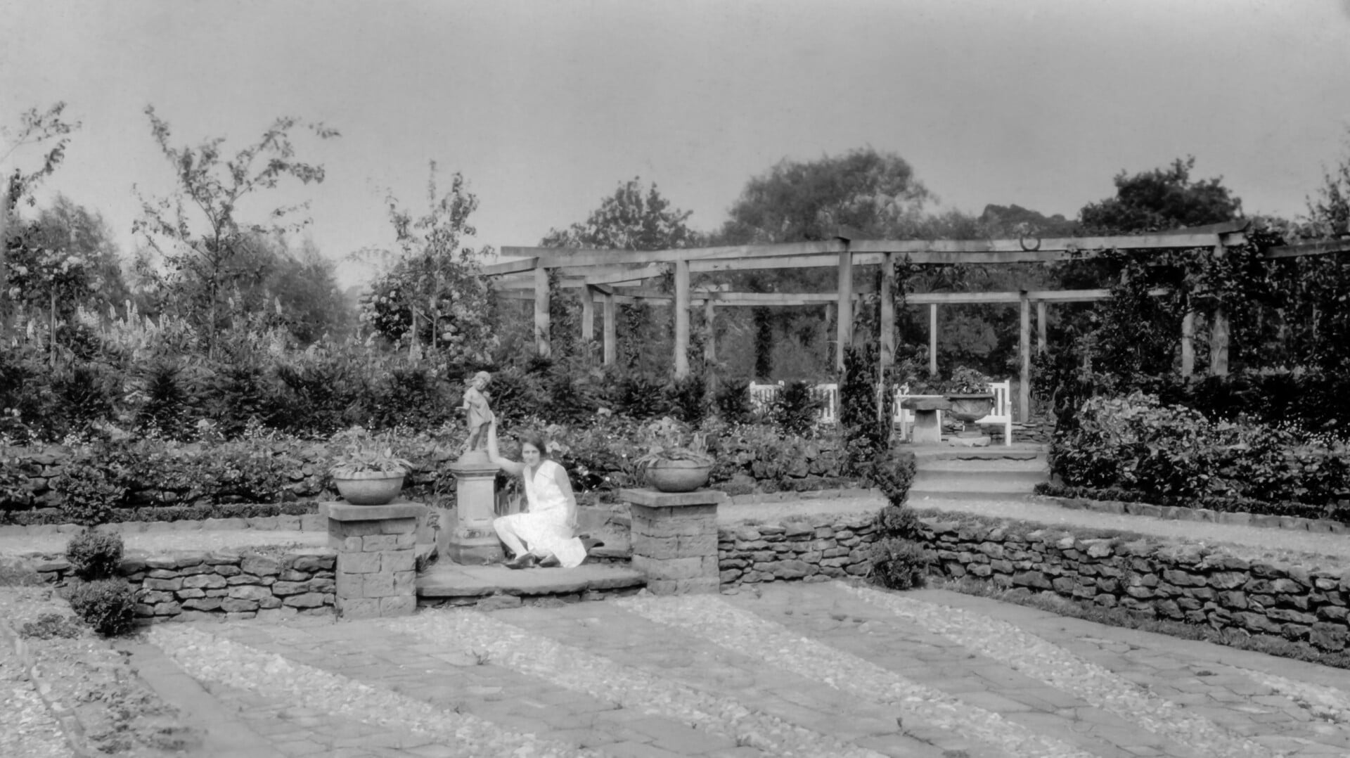 Black and white photo of a woman sitting on a wall in a picturesque garden