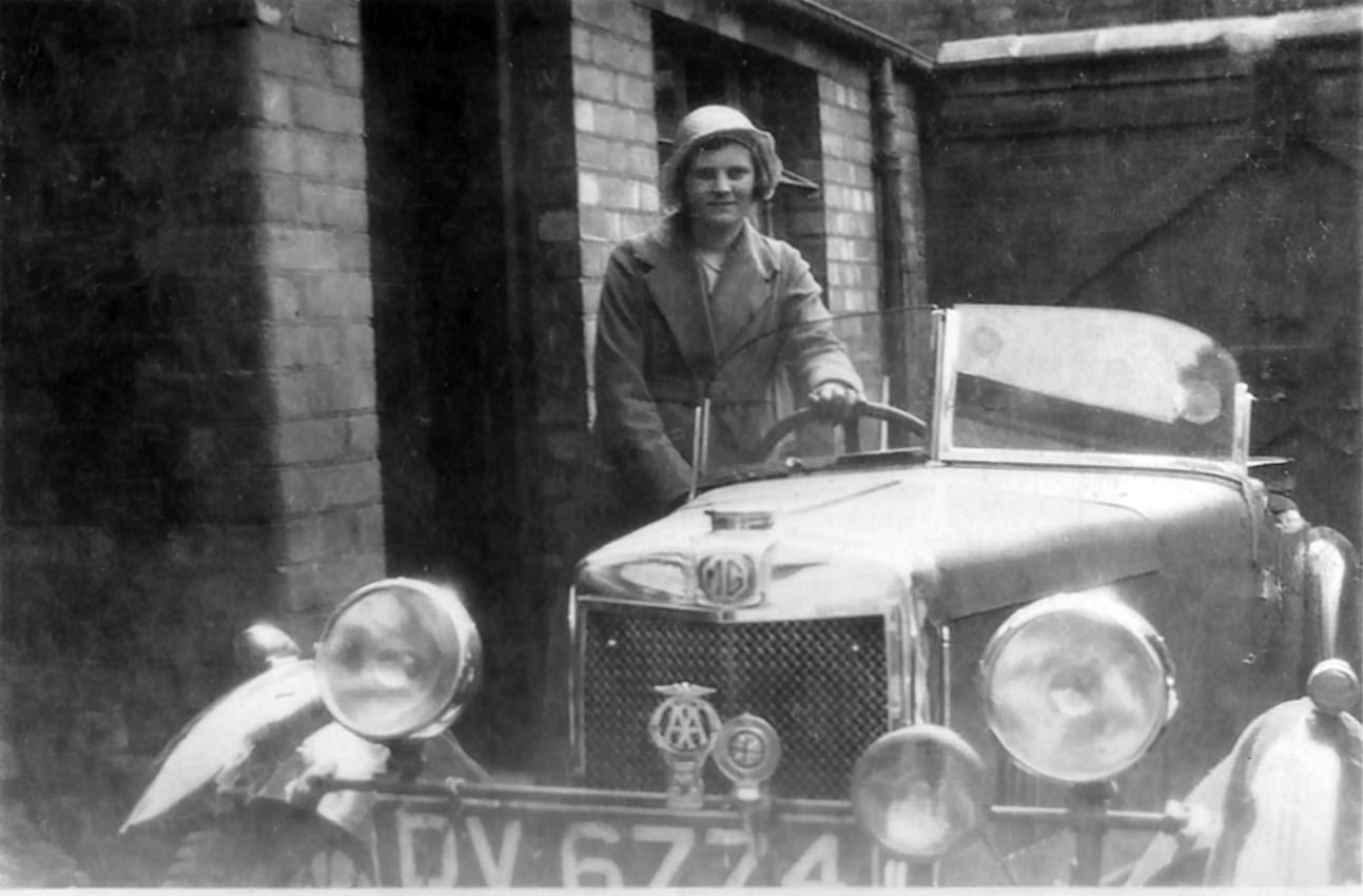 Black and white photo of a woman standing by an old MG motor car