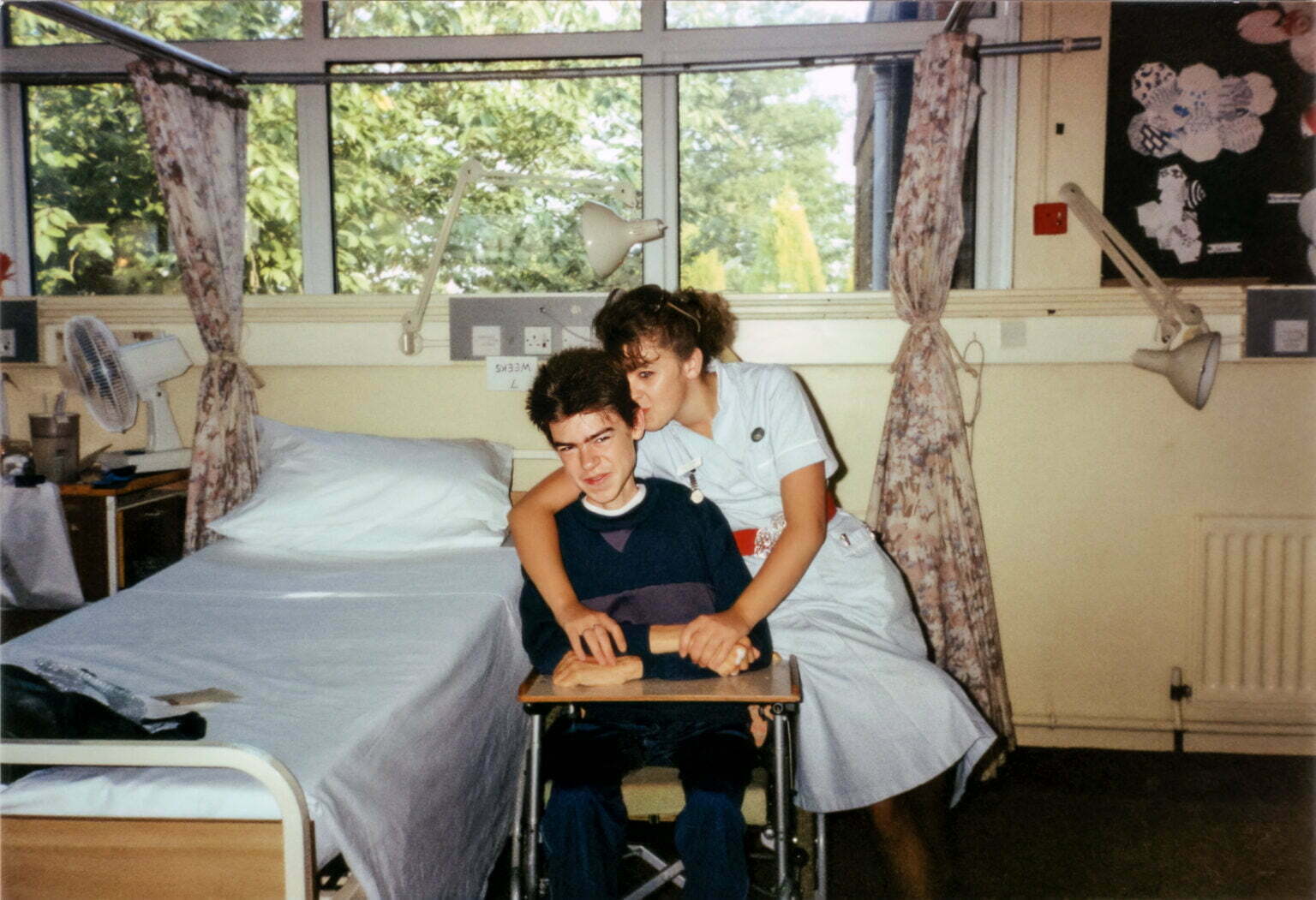 A teenage Daniel Baker in a wheelchair and nurse called Louise hugging him by a hospital bed.