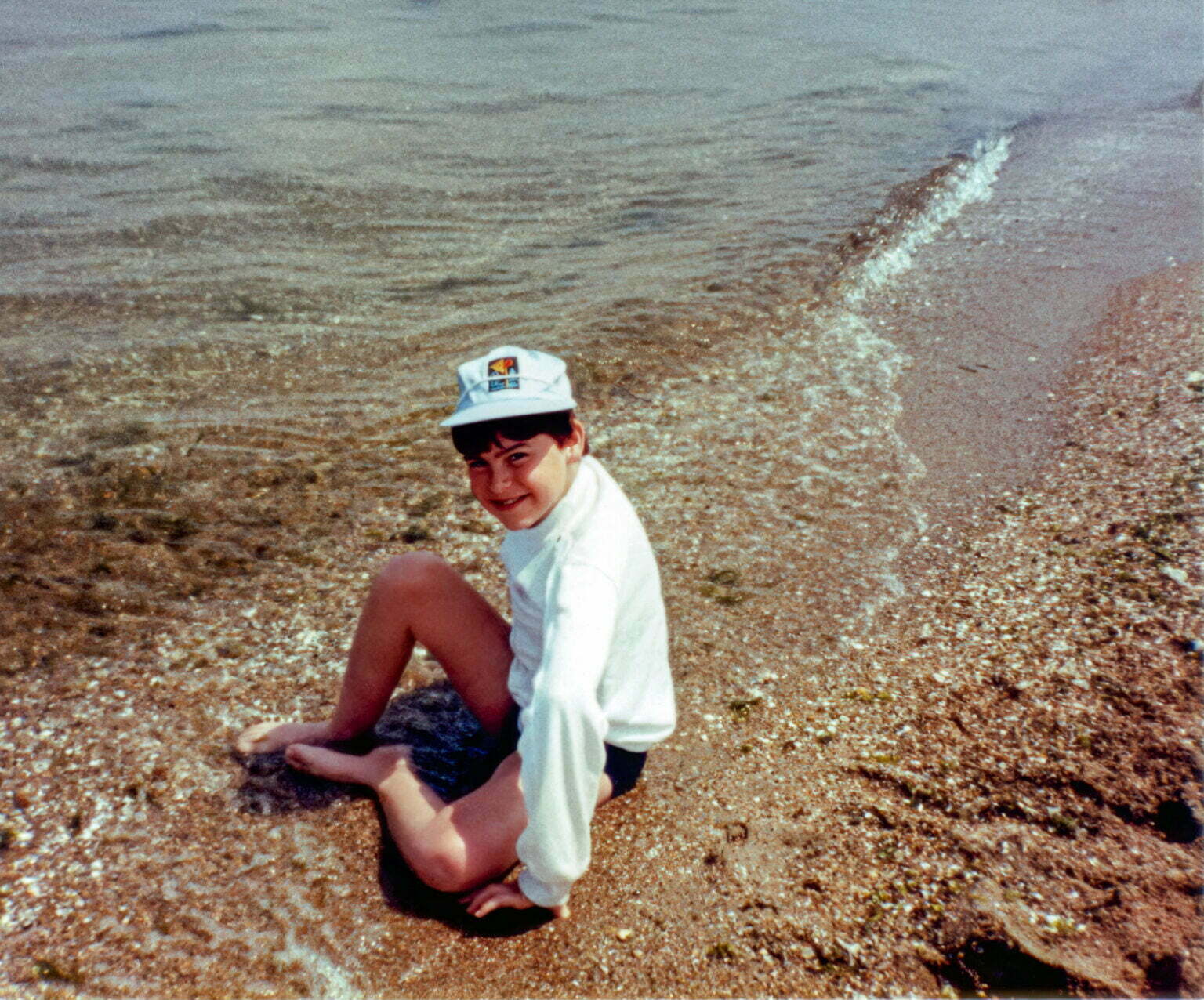 a young boy sitting on a beach next to a body of water in Greece