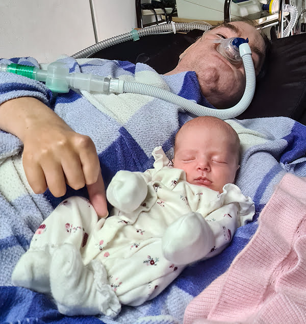 A baby wearing a baby grow laying in the crook of a man's arm