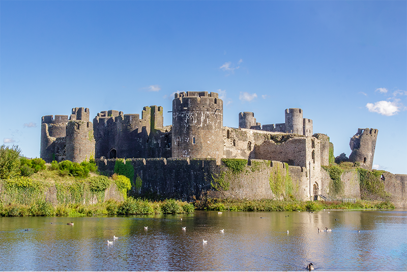 Scenic view of the medieval stone Caerphilly castle with round towers and battlements, reflected in a tranquil moat under a clear blue sky; ivy and greenery hint at its age and historical charm.