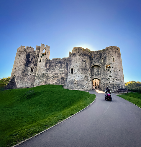 Historic stone Chepstow castle with cylindrical towers silhouetted against a warm sun; a person in a powered wheelchair approaches the arched entrance along a paved path, highlighting the blend of ancient architecture and modern accessibility.
