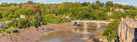 Scenic riverside view with a white arched bridge crossing muddy banks, surrounded by dense greenery and hillside homes; a historic stone castle structure stands on the right, blending natural beauty with architectural heritage.