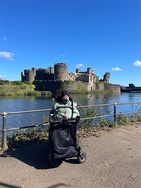 Person in a motorised wheelchair on a moat side path, gazing across the water at a historic stone castle with towers and battlements; the scene is framed by a railing, foliage, and a bright blue sky with scattered clouds, highlighting inclusion in a heritage setting.
