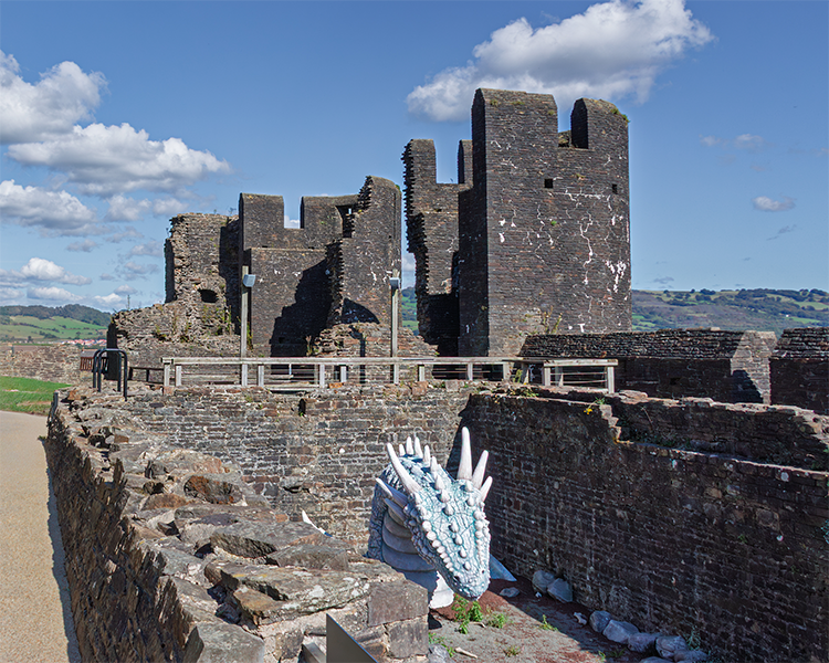 Ruins of historic stone castle with tall rectangular towers, set against green hills and a partly cloudy sky; a striking blue-and-white dragon head sculpture emerges from the ground near the castle wall, blending fantasy with heritage.