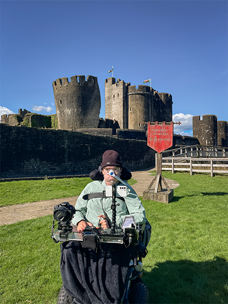 Person in a motorised wheelchair facing a medieval castle with round towers and battlements; equipped with cameras and communication devices, they sit on a stone path beside a red sign warning visitors not to climb the walls. The scene blends historical architecture with modern accessibility under a clear blue sky.