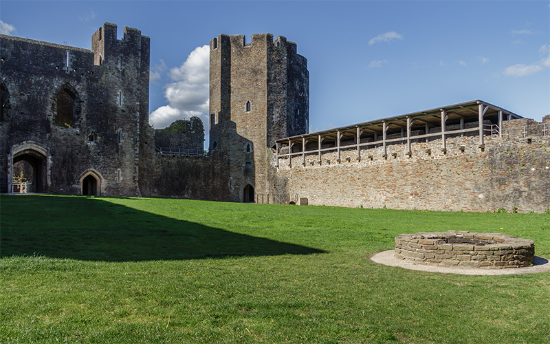 Medieval castle courtyard with two large cylindrical stone towers and a covered gallery along the outer wall; a circular stone well sits on a neatly trimmed lawn under a bright blue sky with scattered clouds, evoking historical architecture and serene atmosphere.