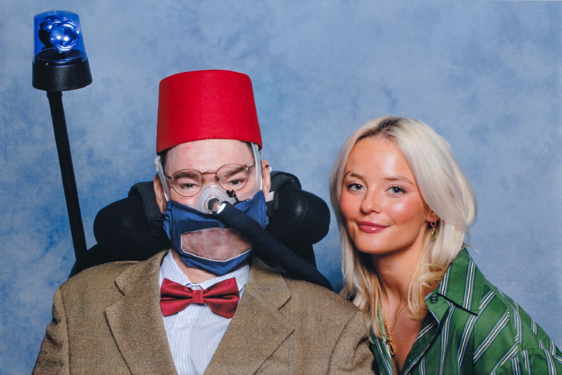 Two people pose together in front of a blue, cloud-patterned backdrop. The person on the left, Daniel Baker, sits in a motorised wheelchair, wearing a red fez, glasses, tweed jacket, red bow tie, and a transparent face mask connected to a ventilator. A blue emergency light is mounted behind the chair. The person on the right, Millie Gibson, stands close, with long blonde hair and a green-and-white striped shirt.