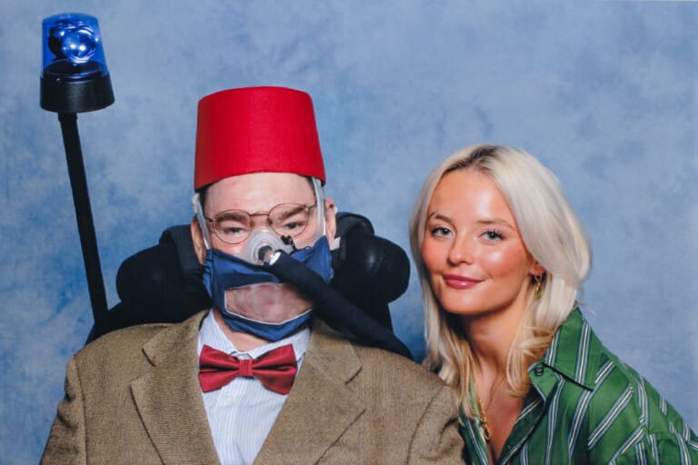 Two people pose together in front of a blue, cloud-patterned backdrop. The person on the left, Daniel Baker, sits in a motorised wheelchair, wearing a red fez, glasses, tweed jacket, red bow tie, and a transparent face mask connected to a ventilator. A blue emergency light is mounted behind the chair. The person on the right, Millie Gibson, stands close, with long blonde hair and a green-and-white striped shirt.