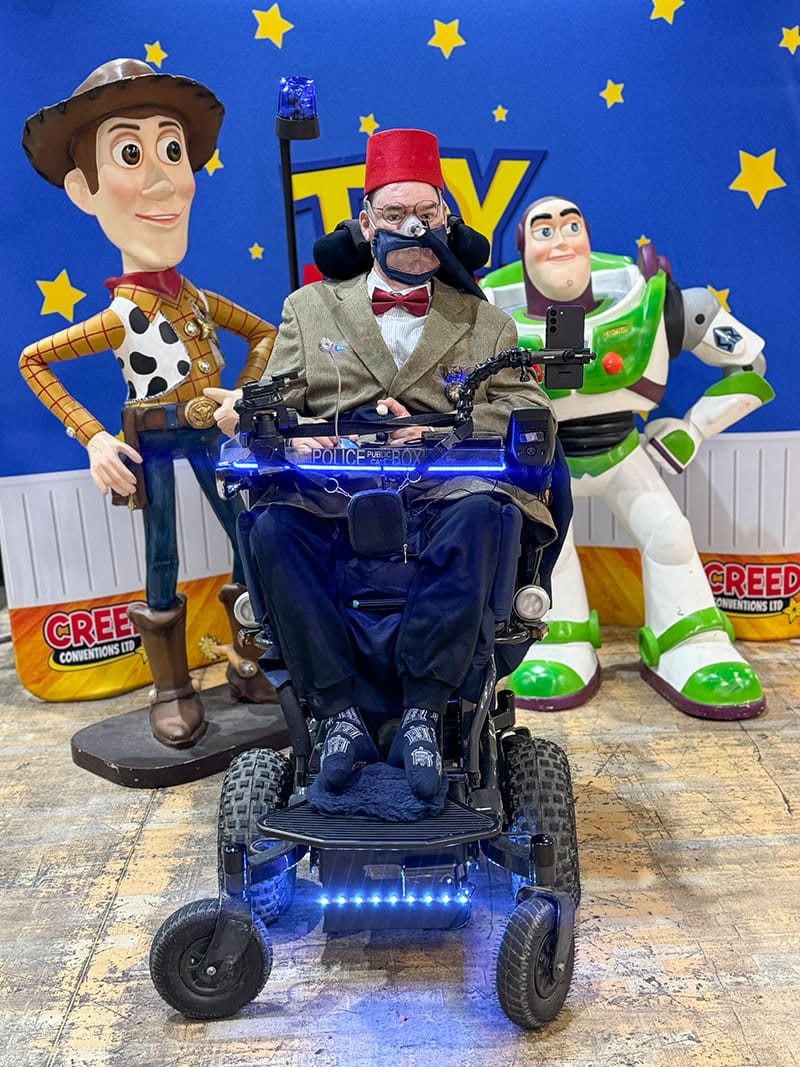 Birthday at Cardiff Comiccon. Daniel Baker in a motorised wheelchair sits between life-sized statues of Woody and Buzz Lightyear. Daniel is wearing a red fez, tweed blazer, and bow tie, with medical equipment and blue LED lights on the chair. A “POLICE BOX” sign is mounted on the front. Behind them, a blue wall with yellow stars and the word “TOY” sets a playful, themed backdrop.