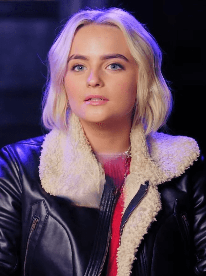 Millie Gibson as Ruby Sunday. A person with short, wavy blonde hair wears a black leather jacket with shearling lining over a red top. They look slightly off-camera with a neutral expression. The dark, softly lit background suggests an indoor or studio setting. This file comes from the official Doctor Who YouTube channel, which has released a limited number of clips under the CC BY licence.