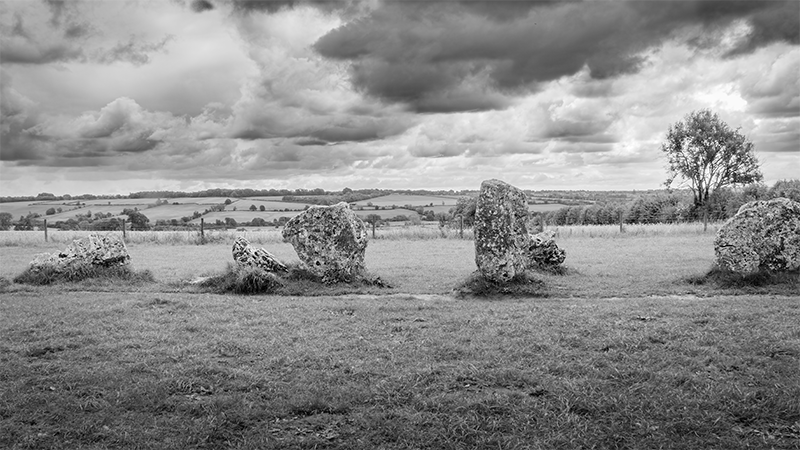 Black and white photograph of a windswept rural landscape featuring a line of ancient standing stones, The Rollright Stones, in the foreground. The weathered, irregular stones rise from a grassy field, backed by rolling countryside and dramatic, cloud-laden skies.