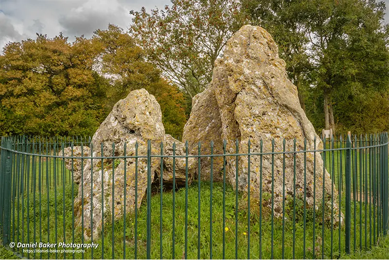 Cluster of large, weathered standing stones enclosed by a metal fence, The Whispering Knights. The irregular stones suggest ancient origins, framed by leafy trees and soft natural light.
