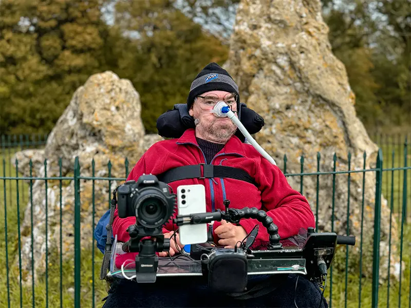 Daniel Baker in a wheelchair wearing a red jacket and black beanie, positioned near ancient standing stones behind a metal fence. The chair is equipped with assistive tech including a breathing tube, smartphone, and mounted camera, suggesting active engagement in media or documentation