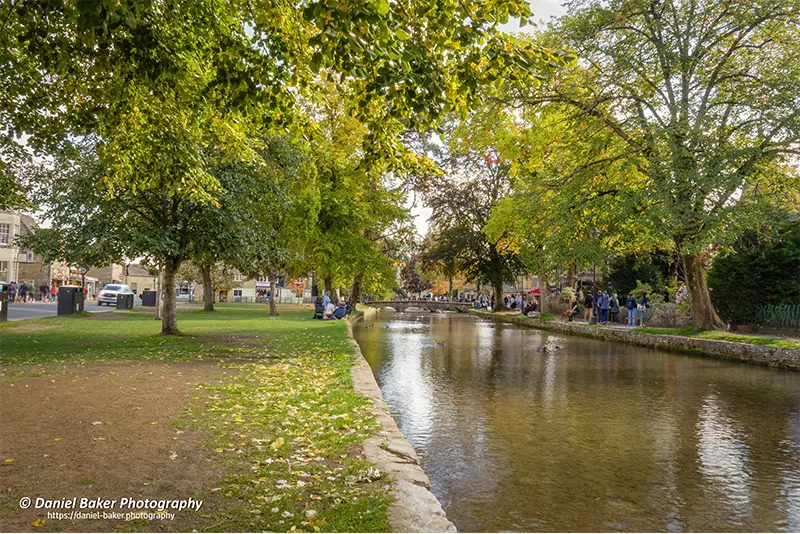 a shallow stream reflecting autumnal trees and sky. People stroll and relax along grassy banks, framed by golden foliage and distant buildings.
