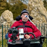Daniel Baker in a wheelchair wearing a red jacket and black beanie, positioned near ancient standing stones behind a metal fence. The chair is equipped with assistive tech including a breathing tube, smartphone, and mounted camera, suggesting active engagement in media or documentation