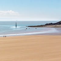 Barry Island. A wide sandy beach curves gently toward grassy dunes under a mostly clear sky. A few people walk along the shoreline, while a tall sculpture stands alone in the shallow water, adding scale and focus to the serene scene.